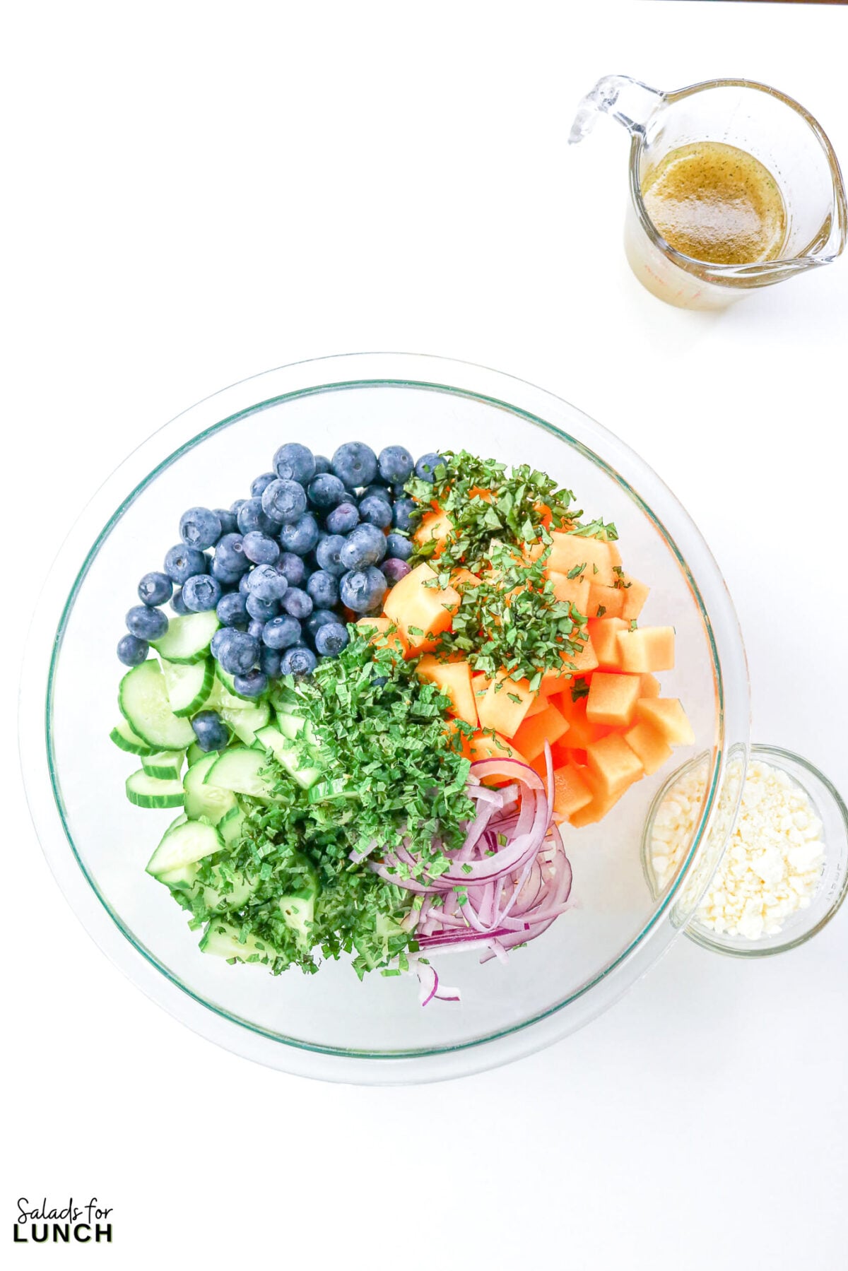 Cantaloupe Cucumber Feta Salad - Cantaloupe Salad Process 5 Bowl of unmixed salad ingredients for cantaloupe cucumber feta salad including blueberries, cucumber, cantaloupe, red onion, and chopped mint, with feta and dressing alongside.