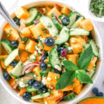 Bowl of cantaloupe salad with cucumber, blueberries, red onion, feta, and mint, garnished with lime.