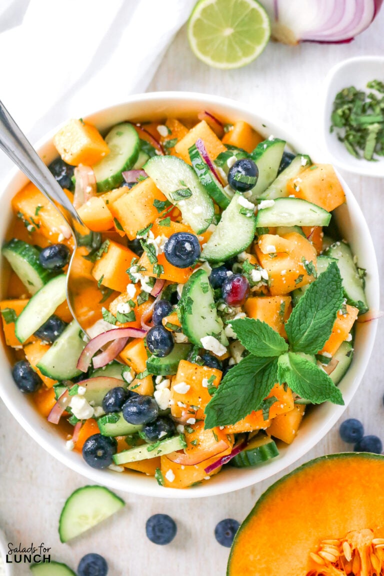 Bowl of cantaloupe salad with cucumber, blueberries, red onion, feta, and mint, garnished with lime.