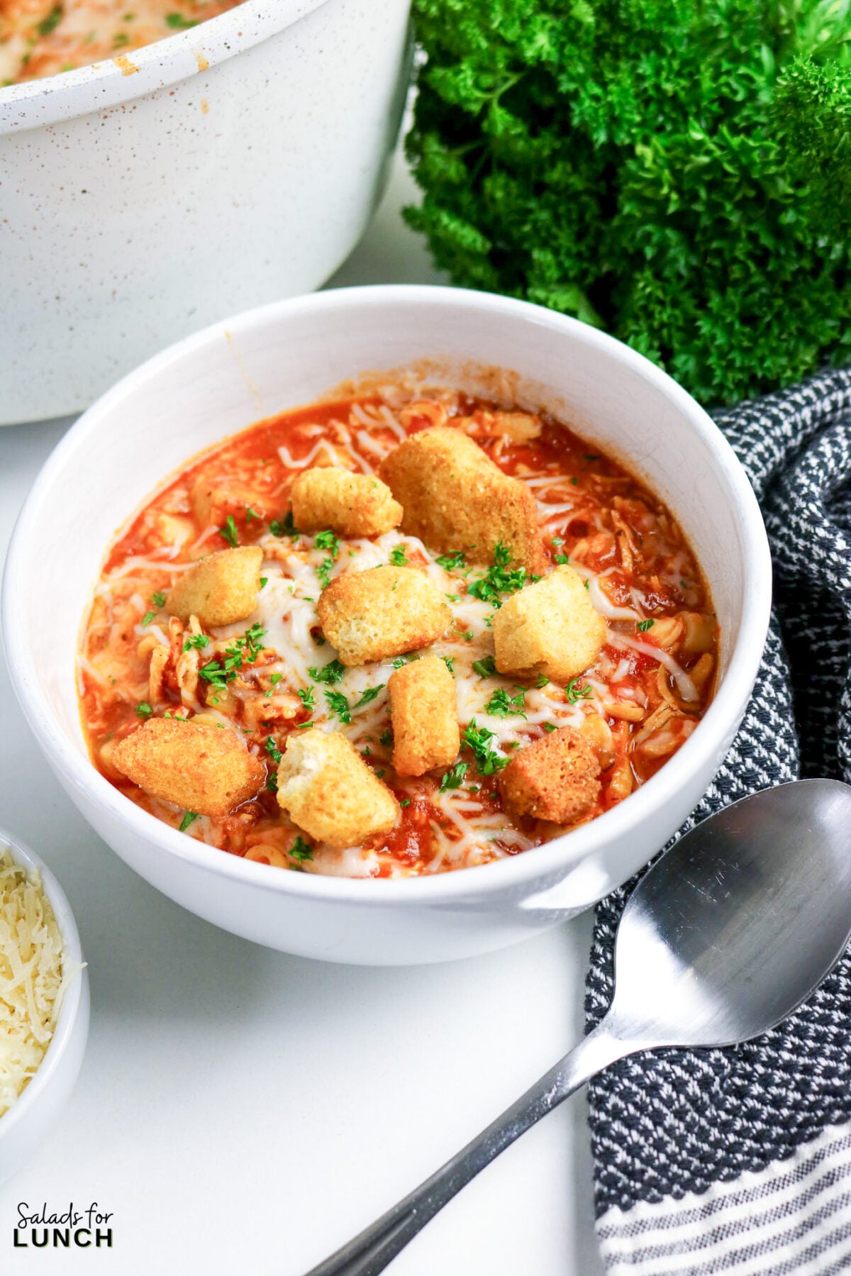 One-Pot Chicken Parmesan Soup - Chicken Parmesan Soup Hero 06 Bowl of tomato-based chicken parmesan soup topped with melted mozzarella, croutons, and chopped parsley, served with a spoon.