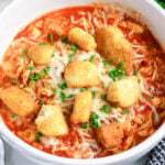 Bowl of tomato-based chicken parm soup topped with melted mozzarella, parsley, and homemade croutons.
