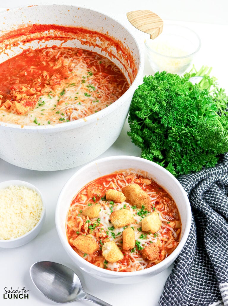 Bowl of Chicken Parmesan soup topped with melted cheese, homemade croutons, and parsley, pot and garnishes nearby.