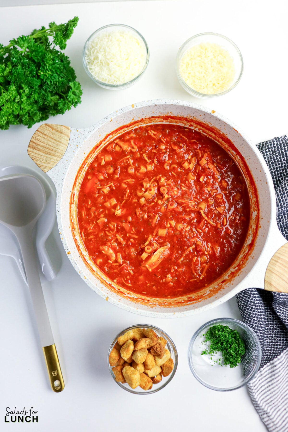One-Pot Chicken Parmesan Soup - Chicken Parmesan Soup Process 07 Pot of chicken parmesan soup with pasta simmered in tomato broth, surrounded by bowls of mozzarella, Parmesan, croutons, and parsley.