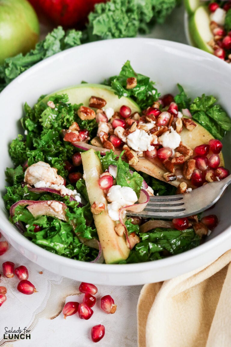 Bowl of kale salad topped with sliced apples, pomegranate seeds, goat cheese, red onion, and chopped pecans.