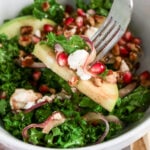 Fork lifting a bite of kale salad with apple slices, pomegranate seeds, goat cheese, pickled red onion, and pecans.