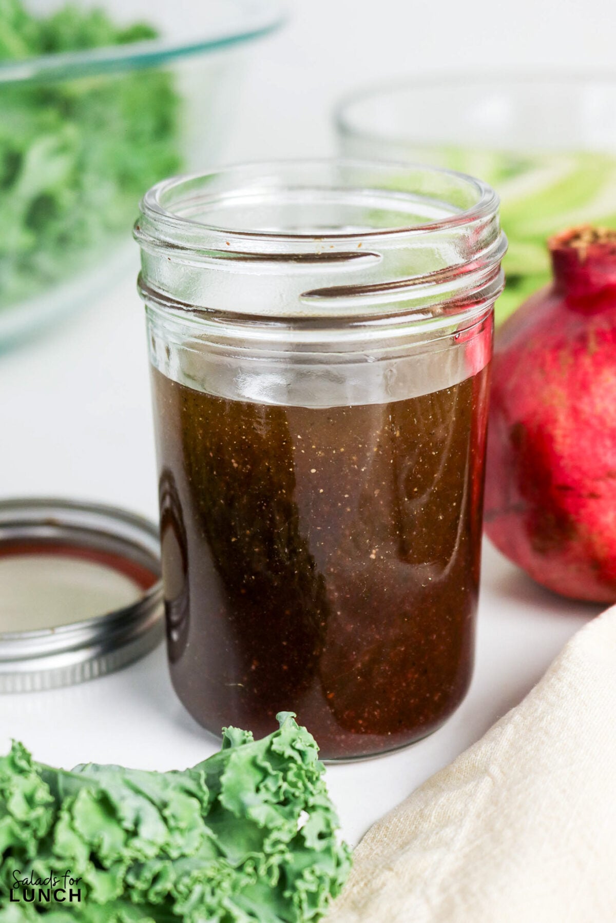 Kale, Apple, Pecan and Pomegranate Salad with Pomegranate Balsamic Dressing - Pomegranate Balsamic Dressing 5 Mason jar filled with dark salad dressing, surrounded by kale leaves and a pomegranate.