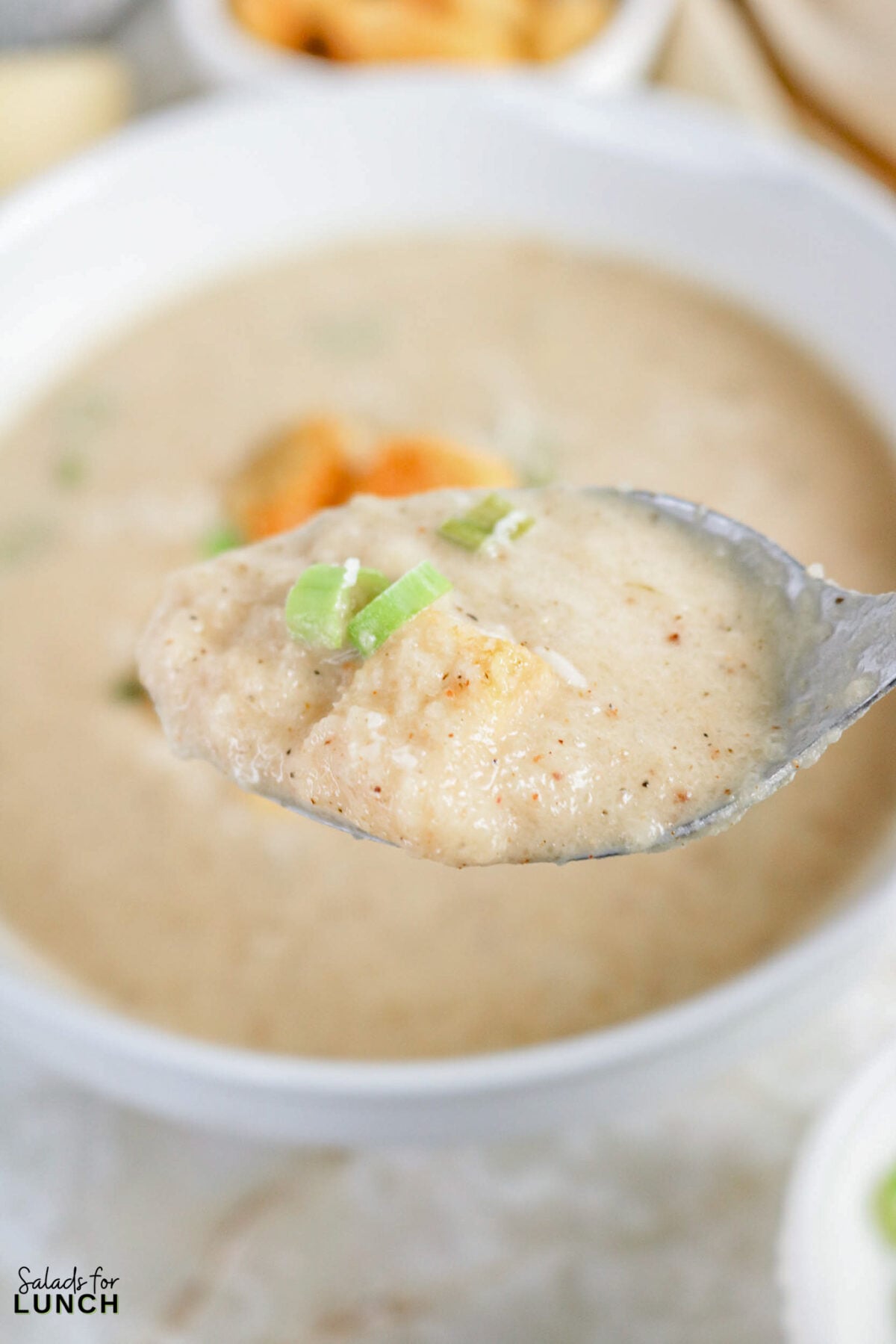 A Spoonful of creamy roasted cauliflower soup with chopped green onion, held above a full bowl of the soup.