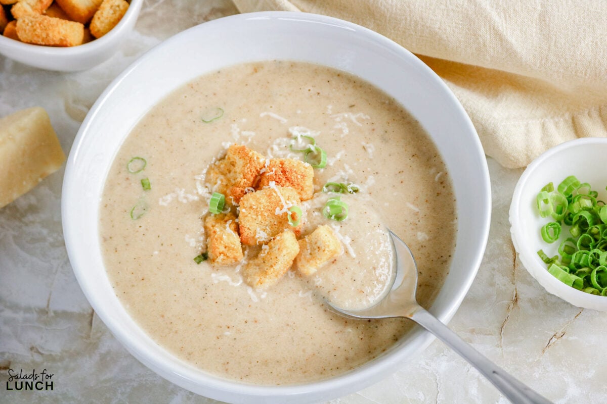 Bowl of creamy roasted cauliflower soup topped with croutons, shredded parmesan, and sliced green onions, spoon resting inside.