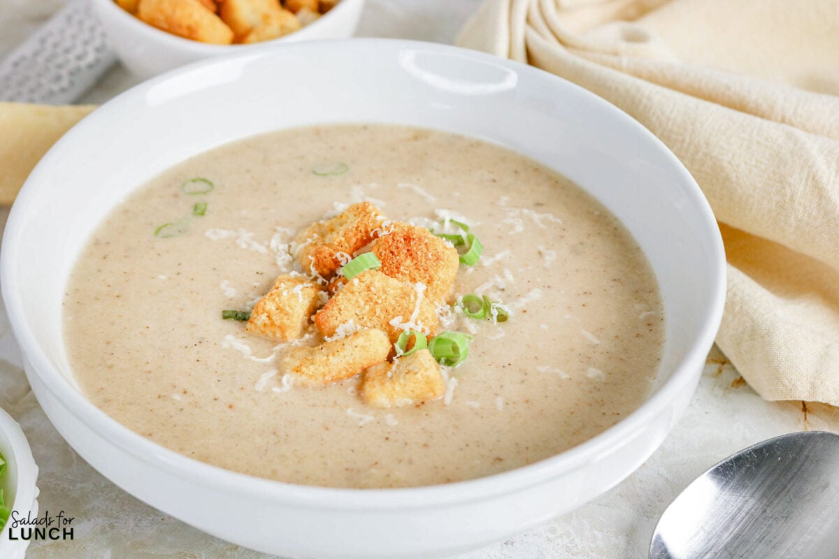 Bowl of creamy roasted cauliflower soup topped with croutons, shredded cheese, and sliced green onions.