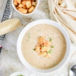Bowl of creamy cauliflower soup topped with croutons, shredded parmesan, and sliced green onions, with extra bowls of croutons and onions alongside a parmesan wedge and grater.