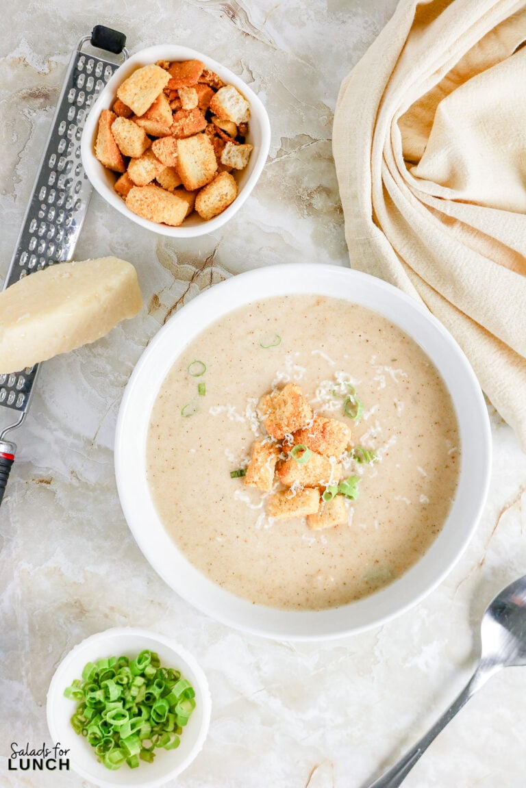 Bowl of creamy cauliflower soup topped with croutons, shredded parmesan, and sliced green onions, with extra bowls of croutons and onions alongside a parmesan wedge and grater.