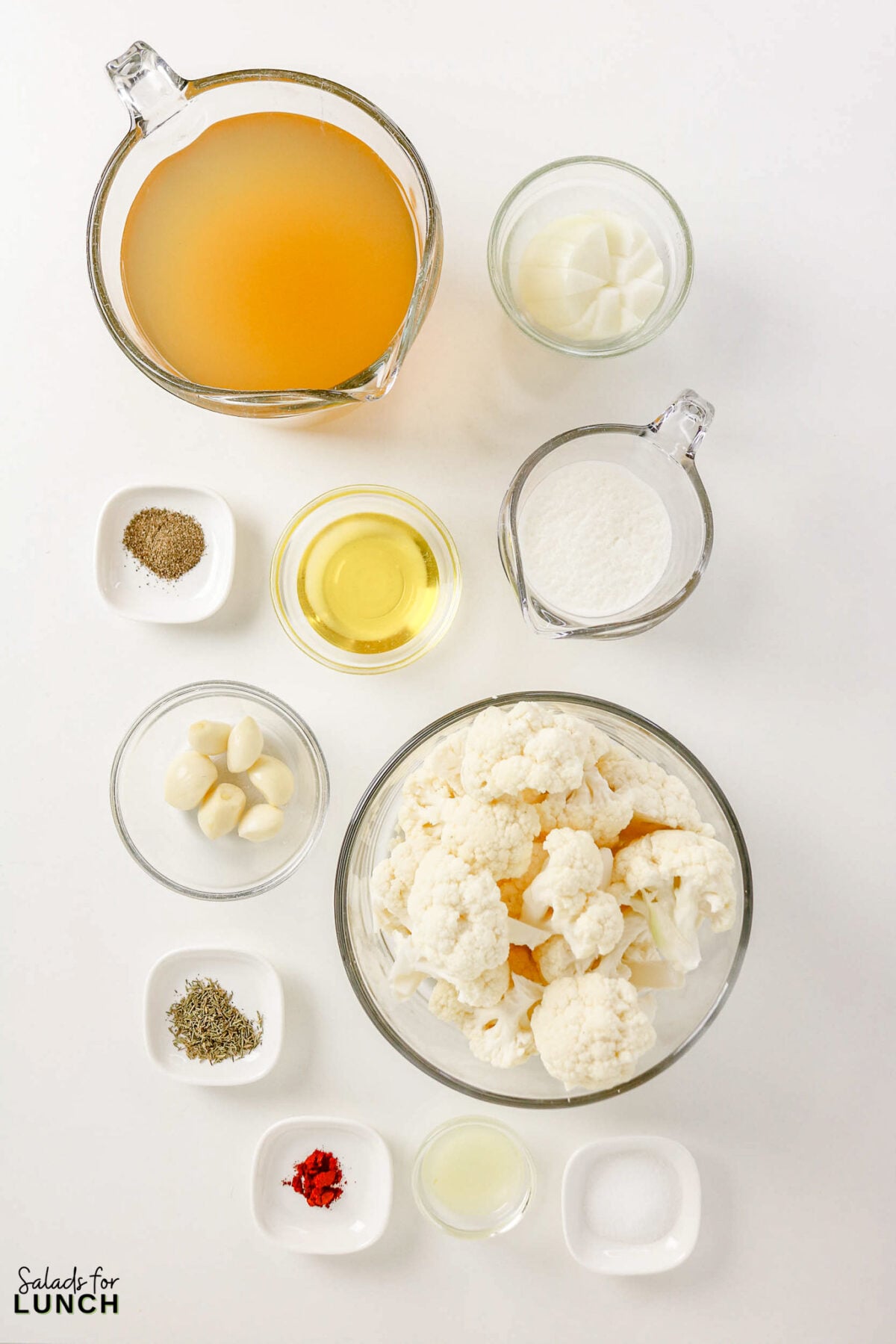 Recipe ingredients for creamy roasted cauliflower soup laid out: broth, cauliflower florets, garlic cloves, oil, flour, paprika, pepper, dried herbs, and lemon juice.