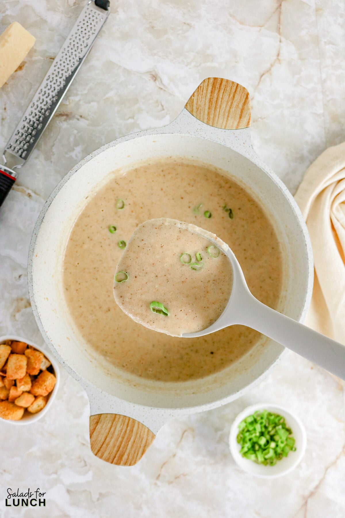 Ladle scooping creamy cauliflower soup topped with sliced green onions from a pot, with bowls of chopped scallions and croutons nearby.