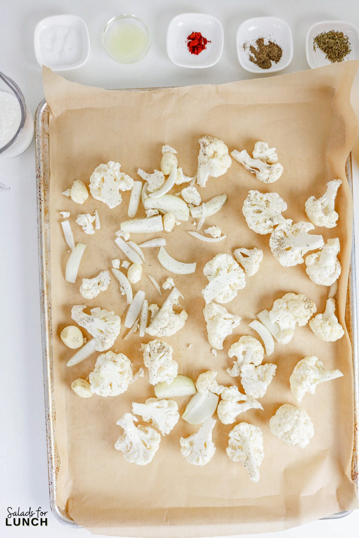 Cauliflower florets, onion, and garlic cloves seasoned and spread on a parchment-lined baking sheet before roasting.