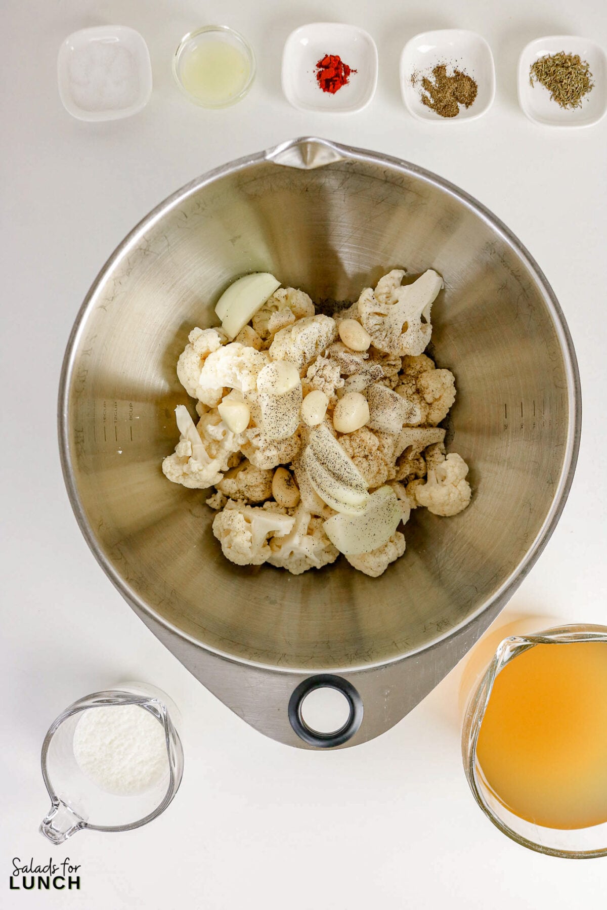 Bowl of cauliflower, onion, and garlic seasoned with oil and pepper, surrounded by broth, cream, and spices before roasting.