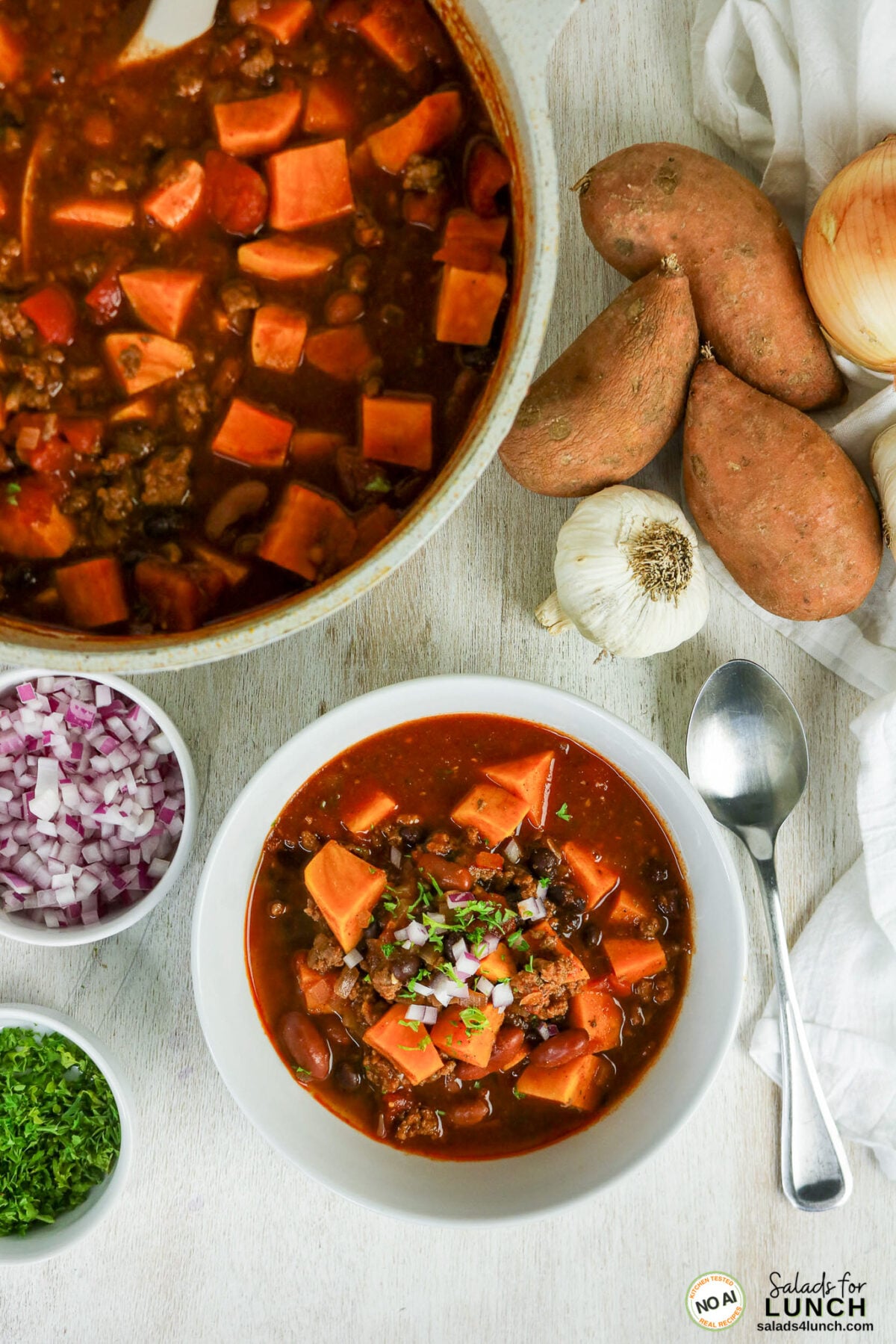 Healthy Sweet Potato Chili with Ground Beef - Sweet Potato Chili Hero 13 Bowl of sweet potato chili topped with red onion and parsley, with pot, sweet potatoes, garlic, and onion toppings nearby.