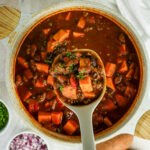 Pot of healthy sweet potato chili with ground beef, black beans, kidney beans, and diced sweet potatoes, with a spoon lifting a scoop, surrounded by diced red onion, chopped parsley, whole garlic, onion, and sweet potatoes.