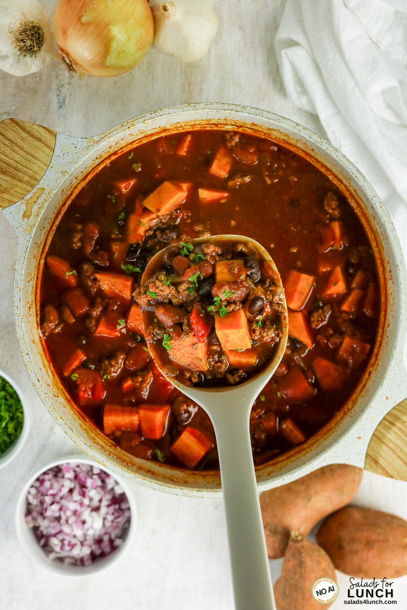 Healthy Sweet Potato Chili with Ground Beef - Sweet Potato Chili Hero 18 Pot of healthy sweet potato chili with ground beef, black beans, kidney beans, and diced sweet potatoes, with a spoon lifting a scoop, surrounded by diced red onion, chopped parsley, whole garlic, onion, and sweet potatoes.