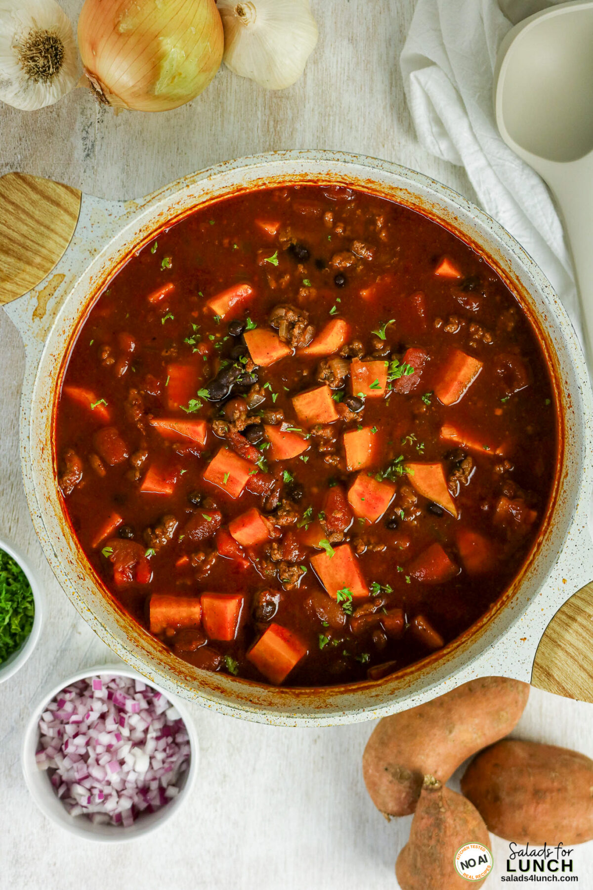 Healthy Sweet Potato Chili with Ground Beef - Sweet Potato Chili Hero 19 Pot of ground beef chili with sweet potato chunks and black beans, surrounded by chopped red onion, cilantro, whole sweet potatoes, onion, and garlic.