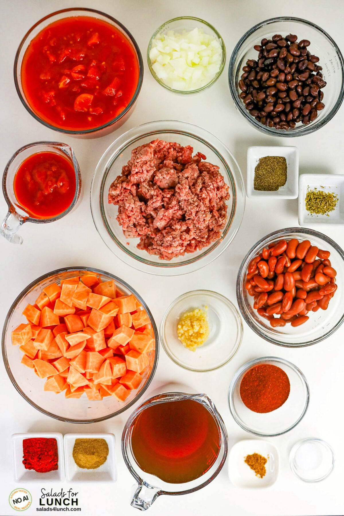 Healthy Sweet Potato Chili with Ground Beef - Sweet Potato Chili Ingredients Overhead view of healthy sweet potato chili ingredients in small bowls: diced tomatoes, onion, black beans, kidney beans, ground meat, cubed sweet potato, minced garlic, broth, and various spices including chili powder, cumin, and paprika.