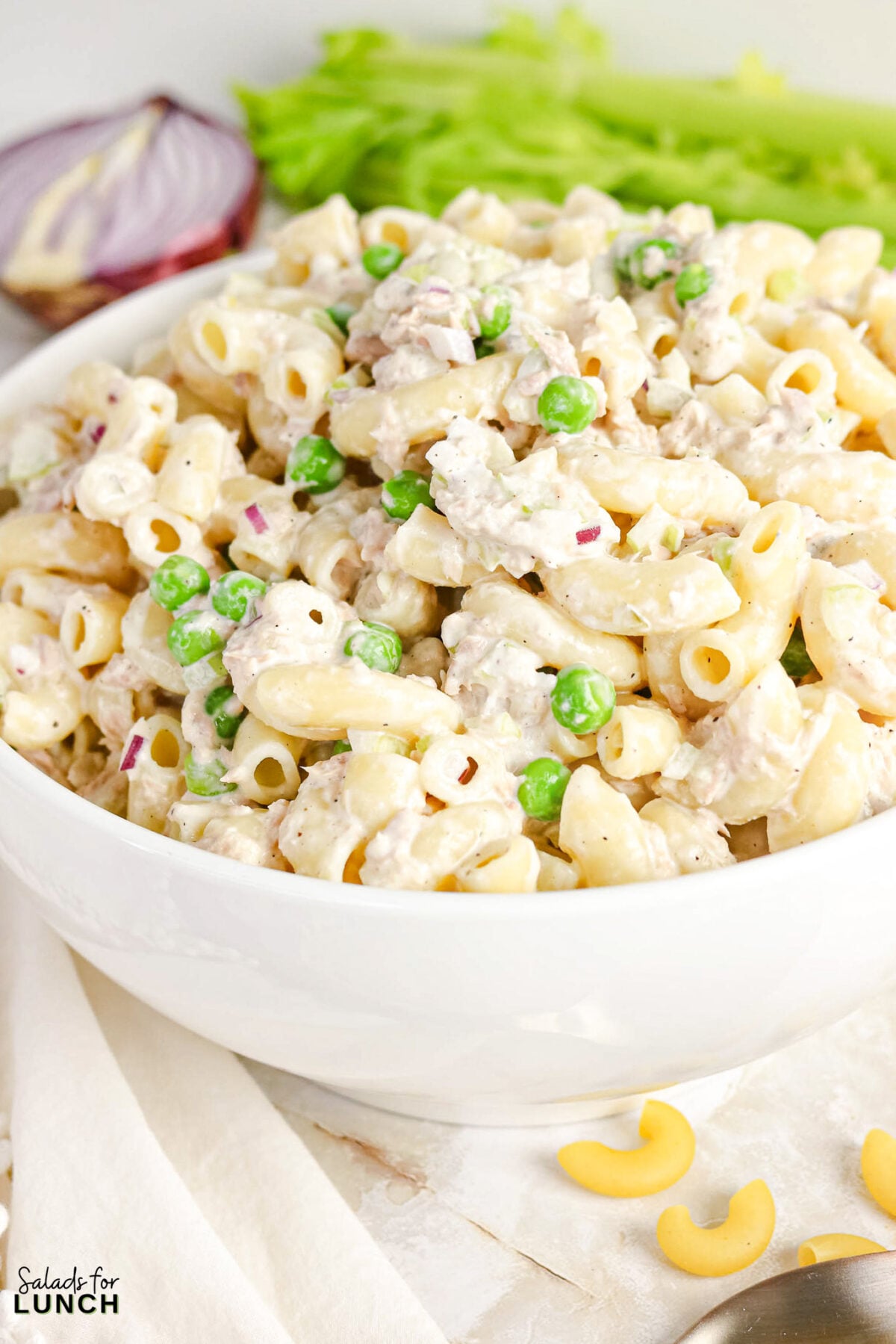 Creamy Tuna Macaroni Salad - Tuna Macaroni Salad Hero 14 Bowl of creamy tuna macaroni salad with peas and diced red onion, garnished with celery.