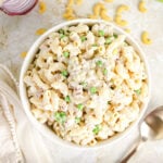 Bowl of creamy tuna macaroni salad with peas, celery, and red onion, surrounded by raw pasta, celery, and a halved red onion.