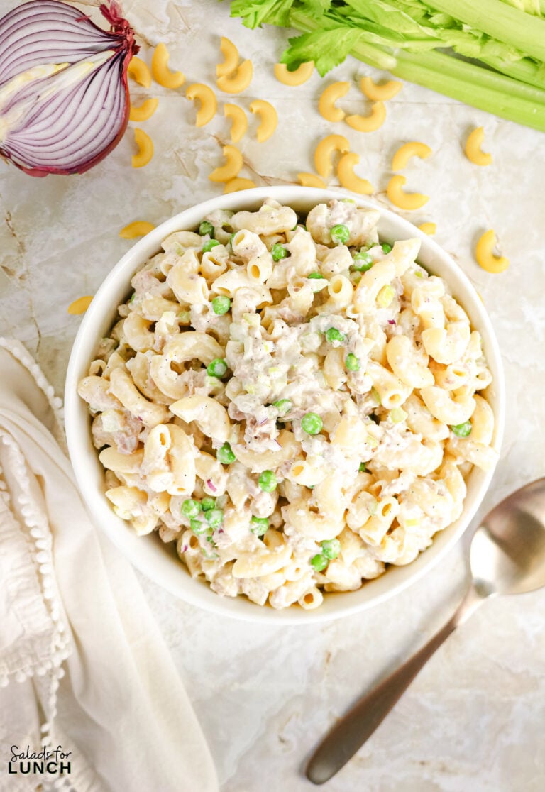 Bowl of creamy tuna macaroni salad with peas, celery, and red onion, surrounded by raw pasta, celery, and a halved red onion.