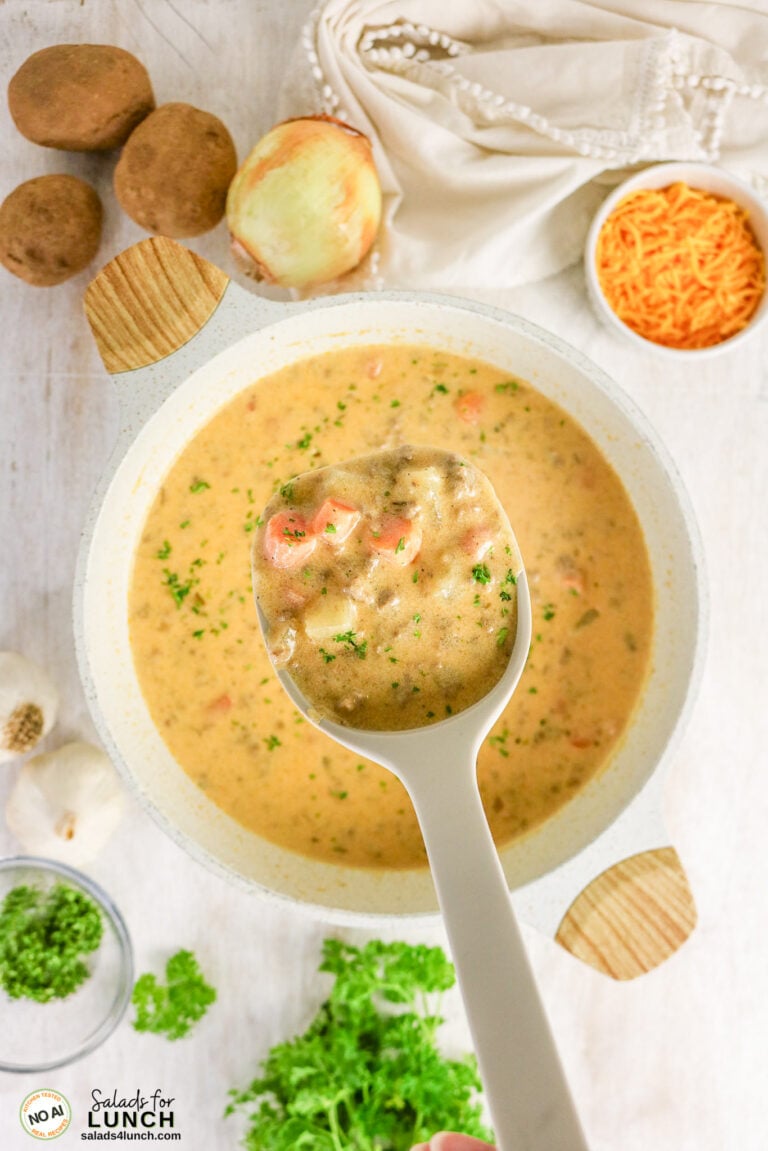 Ladle lifting creamy cheeseburger soup with carrots, surrounded by potatoes, onion, shredded cheese, and parsley.