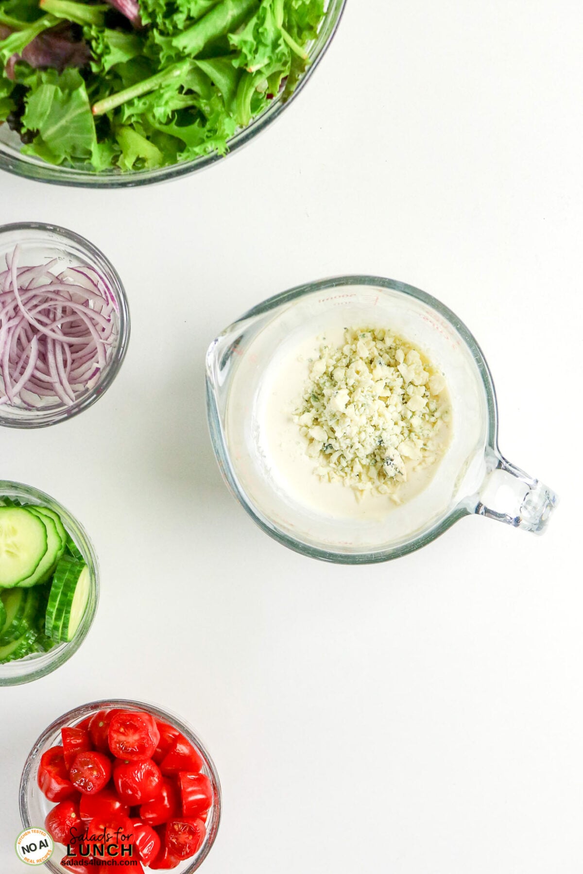 Homemade Blue Cheese Dressing - Homemade Blue Cheese Dressing 1 Overhead view of salad ingredients: mixed greens, sliced red onion, cucumber, cherry tomatoes, and a measuring cup with creamy dressing topped with crumbled blue cheese.