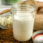 Creamy homemade blue cheese dressing in an open mason jar, with crumbled cheese and salad greens in the background.