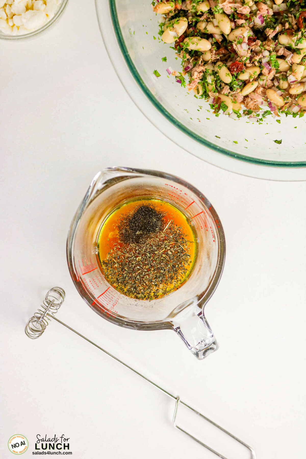 Tuscan Tuna Salad - Tuscan Tuna Salad Process 10 Olive oil, herbs, and spices in a measuring cup beside a mini whisk and bowl of white bean tuna salad.